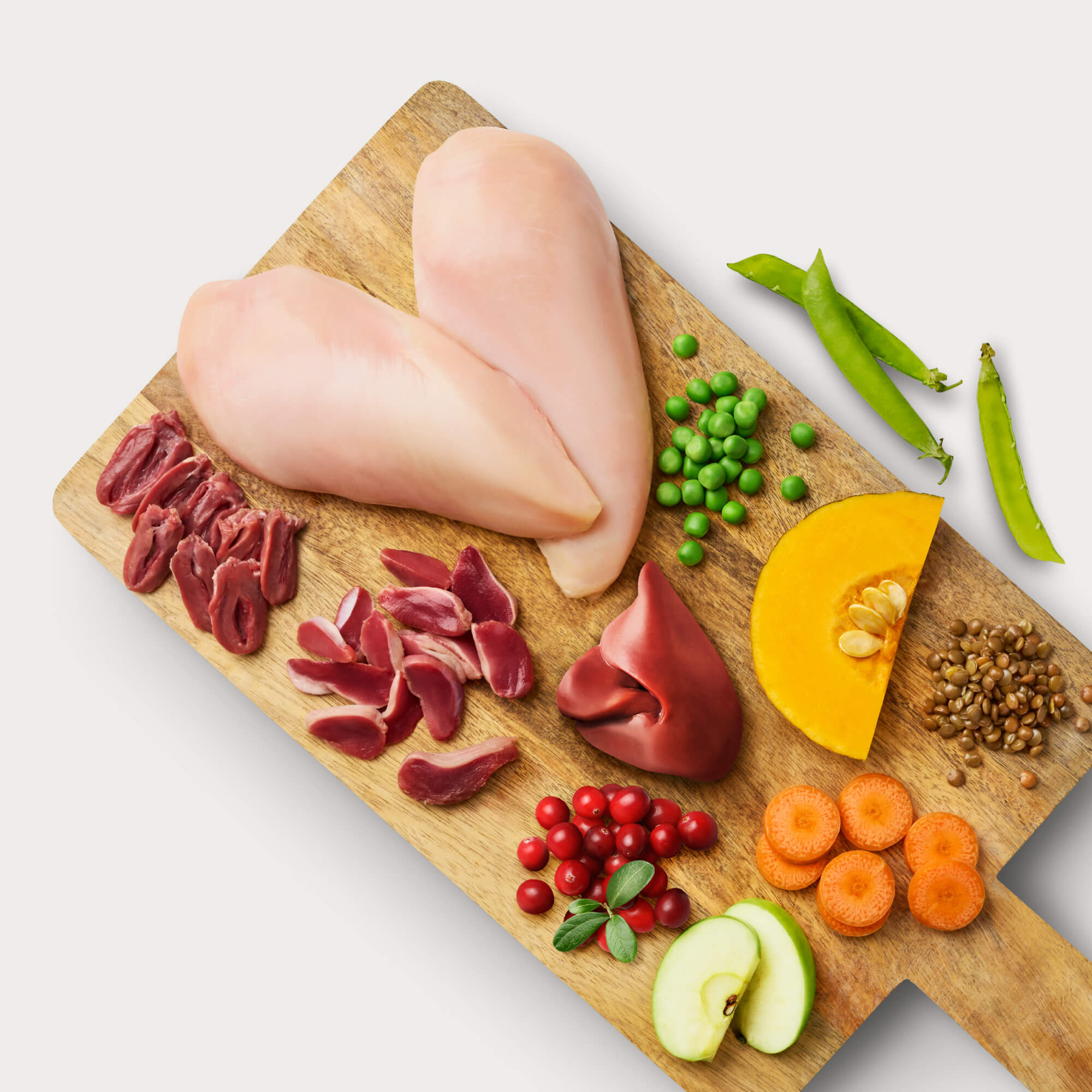 Raw dog food ingredients on wooden cutting board: chicken, chicken organs, lentils, peas, carrots, cranberries, apples, and pumpkin.