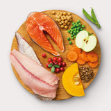 Raw dog food ingredients on wooden cutting board: ocean fish, salmon, lentils, garbanzo beans, peas, carrots, cranberries, apples, and pumpkin.