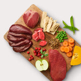 Raw dog food ingredients on wooden cutting board: beef, beef organs, lentils, peas, carrots, cranberries, apples, and pumpkin.
